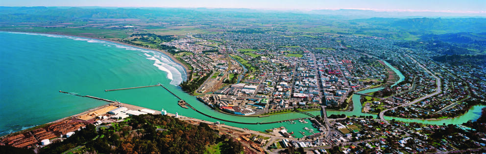 Aerial view of Gisborne, with a river winding through the urban area to the sea, alongside beaches, a harbour and surrounding hills.