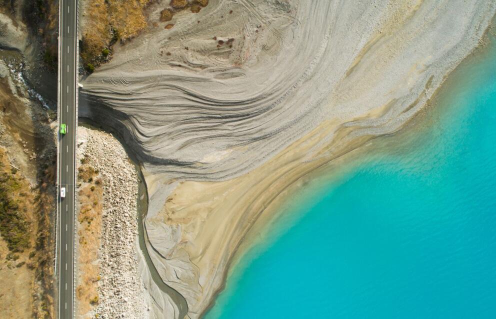 Aerial view of the turquoise waters of Lake Pukaki beside layered glacial sediments and a road running along the shoreline in Canterbury